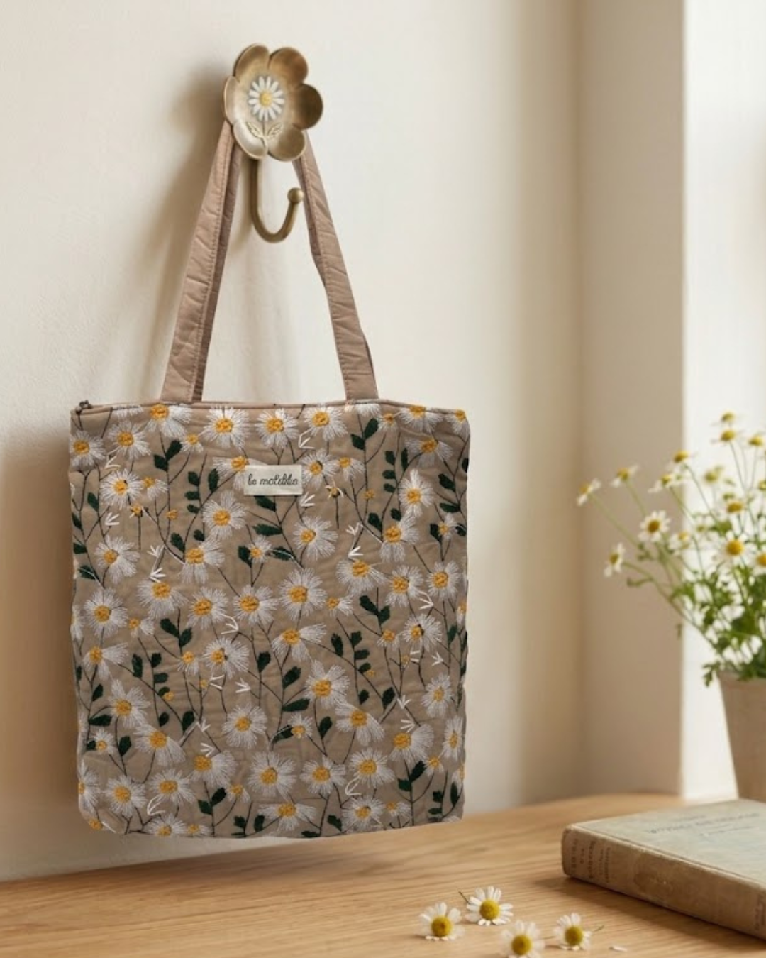 Bolso tote bag acolchado beige con delicado bordado de margaritas blancas y amarillas colgado en percha rústica.