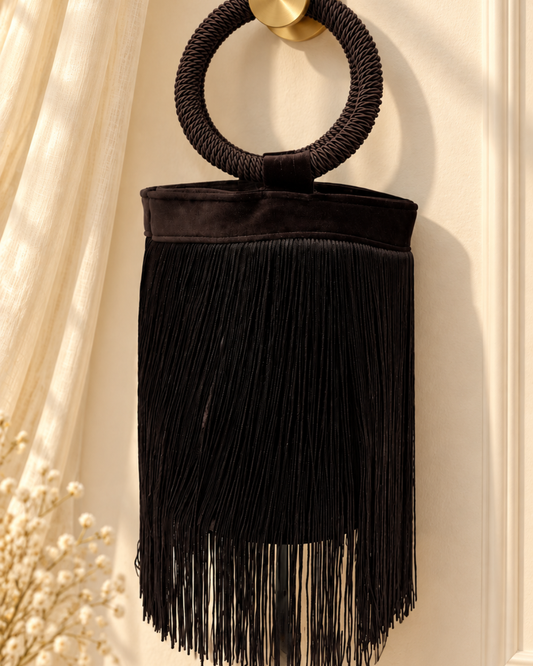 Bolso de terciopelo marrón chocolate muy oscuro con flecos largos y asa circular revestida de cordón, visto de frente sobre fondo beige claro.