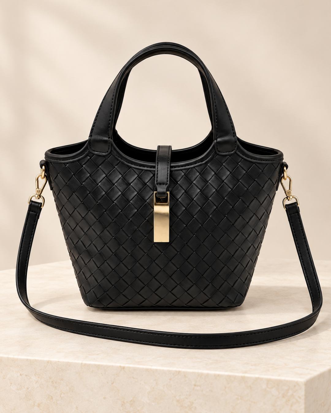 Bolso negro trenzado de mujer con doble asa, detalle rectangular dorado y correa bandolera enganchada a los lados sobre base de piedra clara.