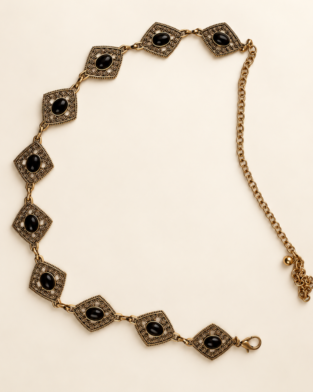 Cinturón joya dorado envejecido con rombos de flores caladas, detalles ovalados negros y cadena ajustable.