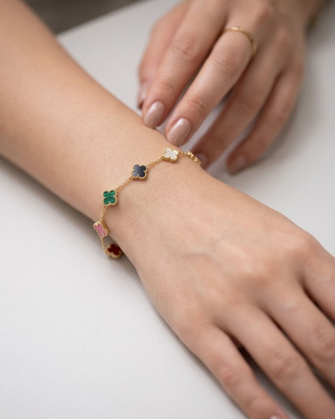 Primer plano de la muñeca de una mujer luciendo una pulsera de cadena de oro fina con cinco dijes de trébol de cuatro hojas de diferentes colores: rojo, rosa, verde malachite, negro y nácar blanco.