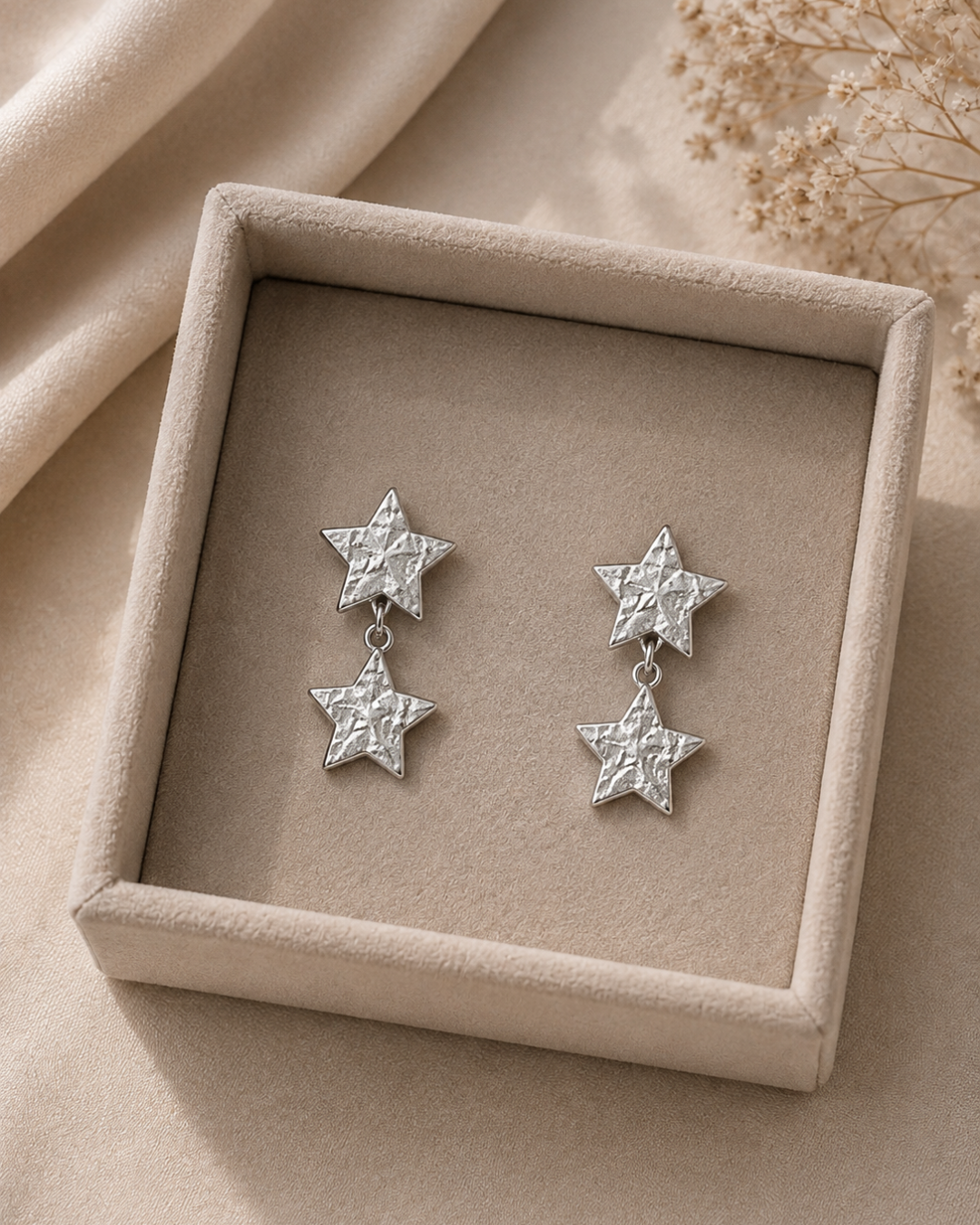 Pendientes de acero inoxidable plateado con forma de doble estrella labrada sobre estuche beige de Accesoriomanía.