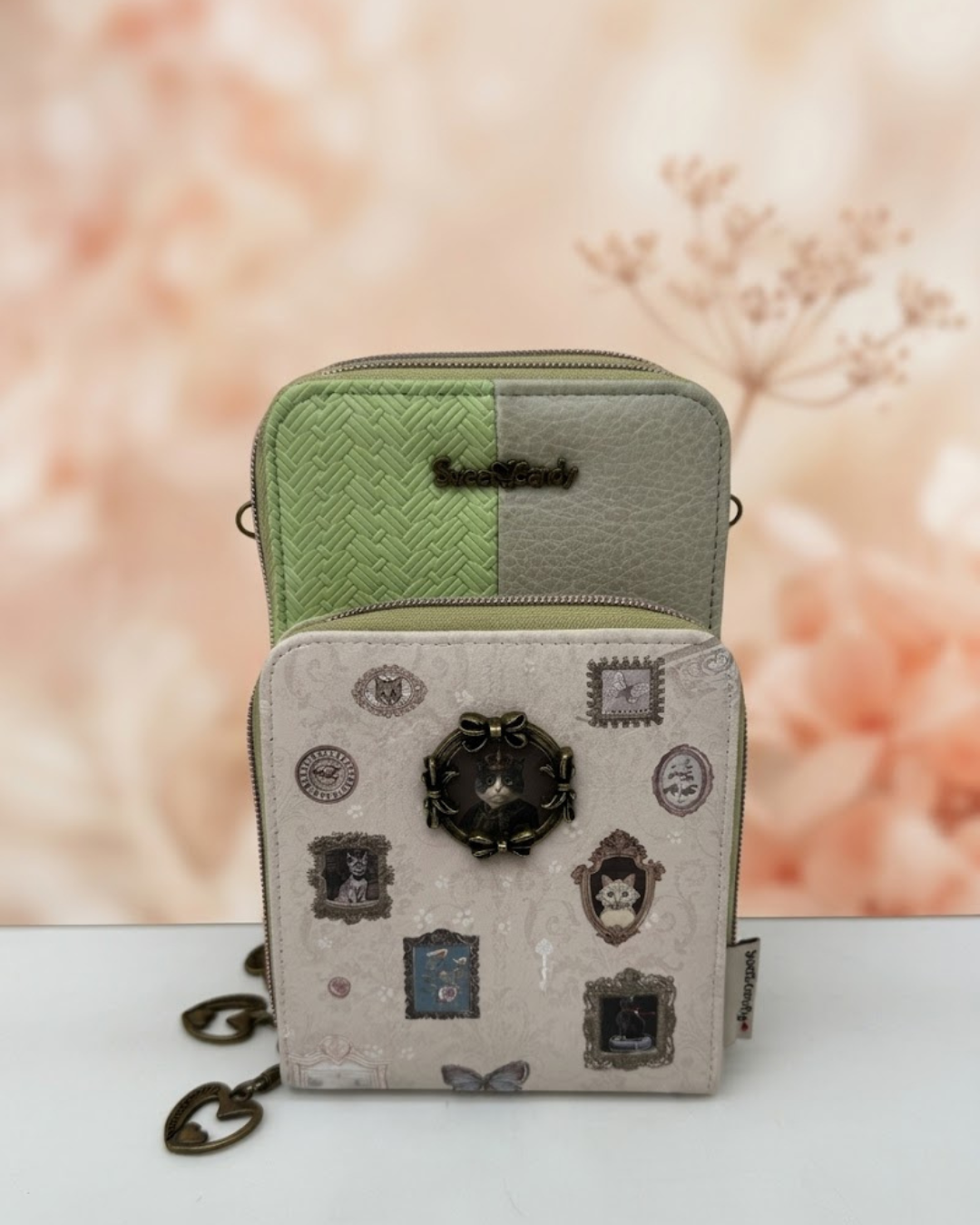 Bolso portamóvil Sweet Candy verde y beige con marco metálico de gatito, billetero y cremalleras sobre fondo floral desenfocado.