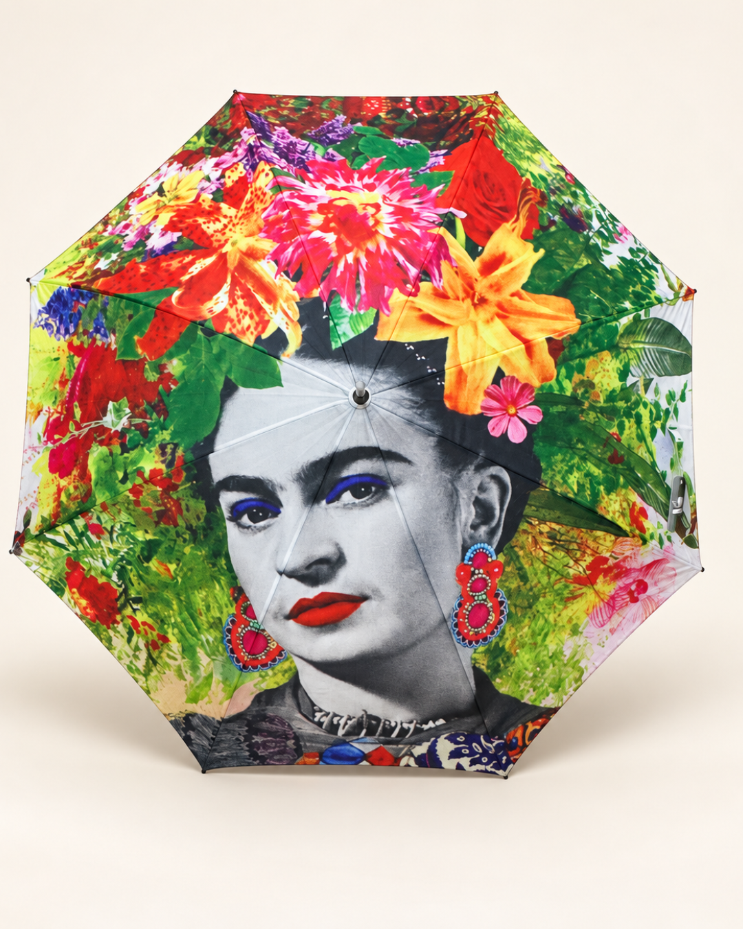 Paraguas abierto con retrato de Frida Kahlo en blanco y negro, flores multicolor y hojas verdes sobre fondo blanco roto.