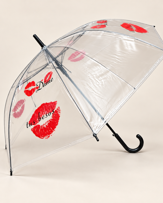 Paraguas transparente abierto de perfil con besos rojos, frase Dame tus besos y mango curvo negro sobre fondo blanco roto.