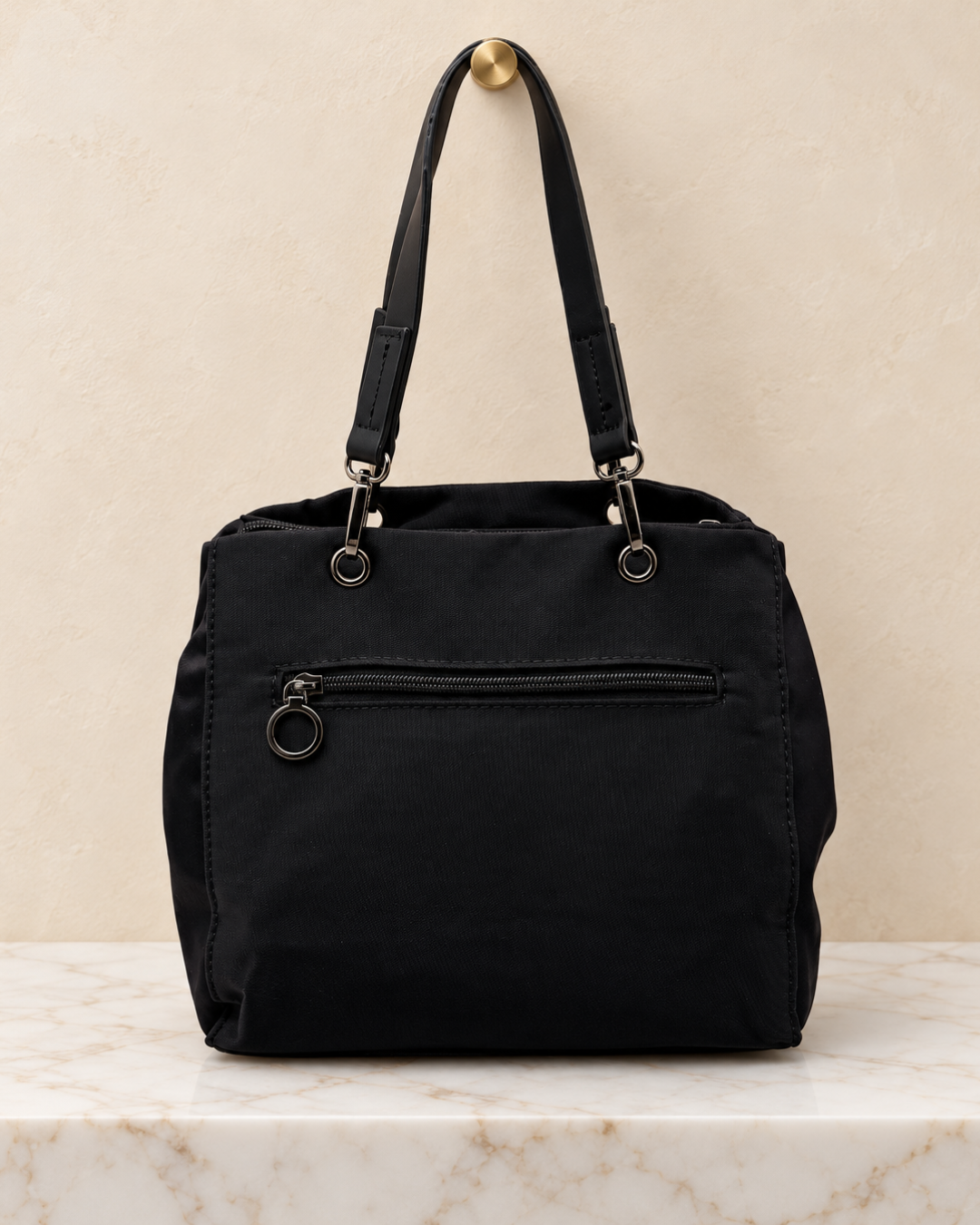 Parte trasera de bolso negro con bolsillo de cremallera y tirador de anilla, apoyado sobre una base de mármol.