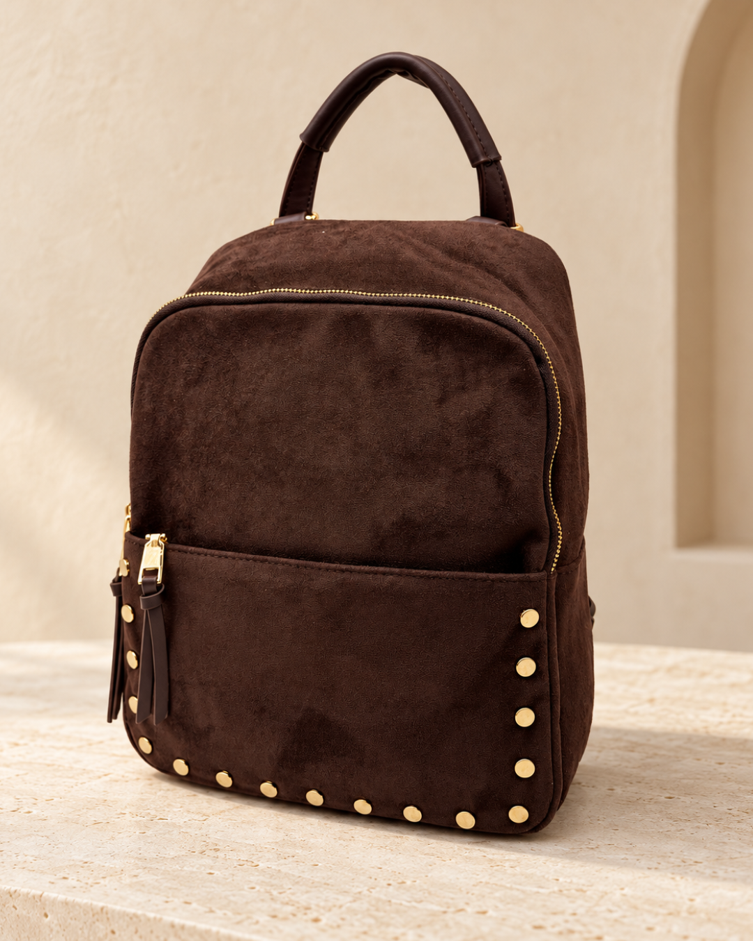 Vista en ángulo de mochila marrón chocolate de efecto antelina con cremalleras doradas, asa superior y tachuelas.