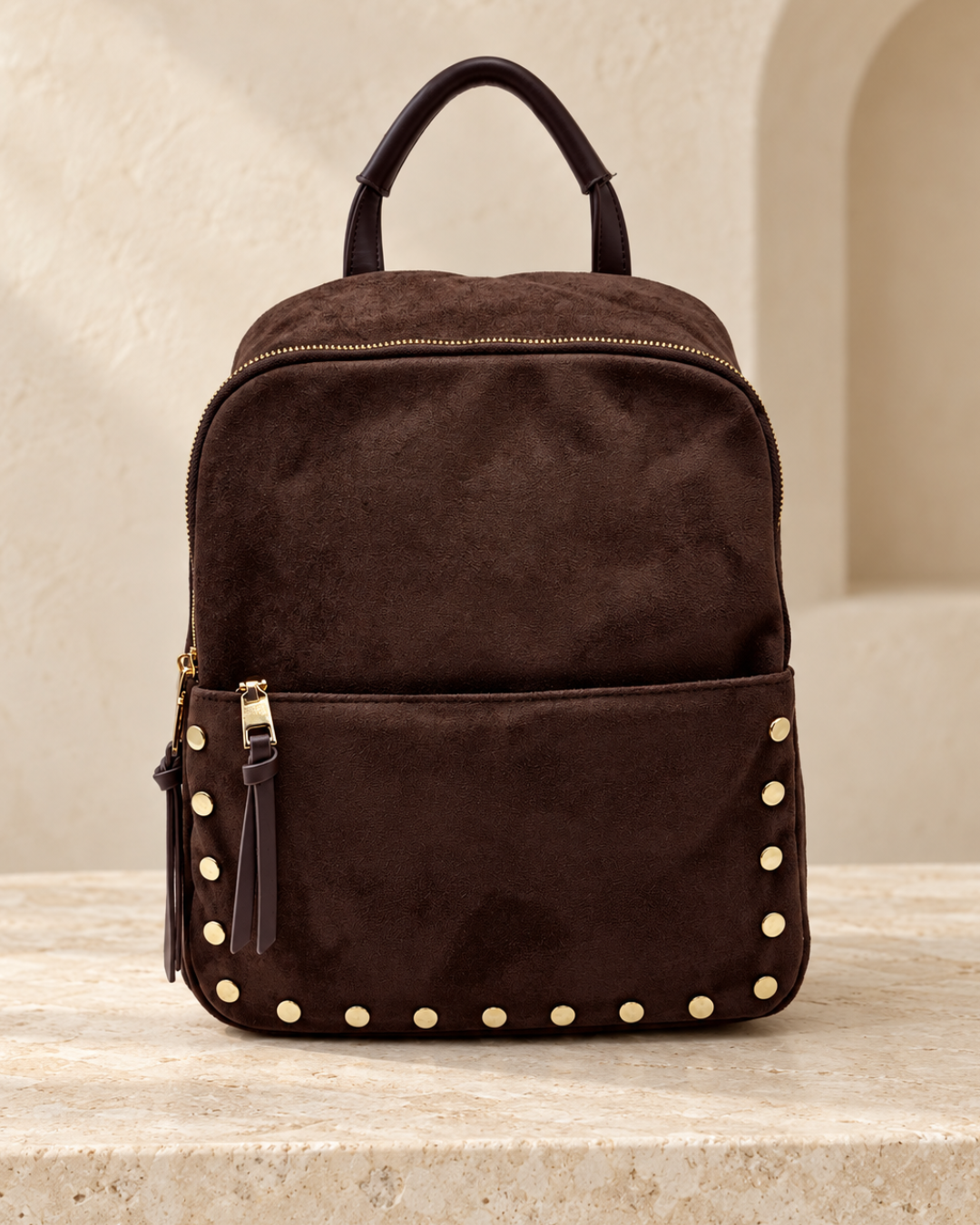Vista frontal de mochila mediana de efecto antelina marrón chocolate con bolsillo delantero y tachuelas doradas.