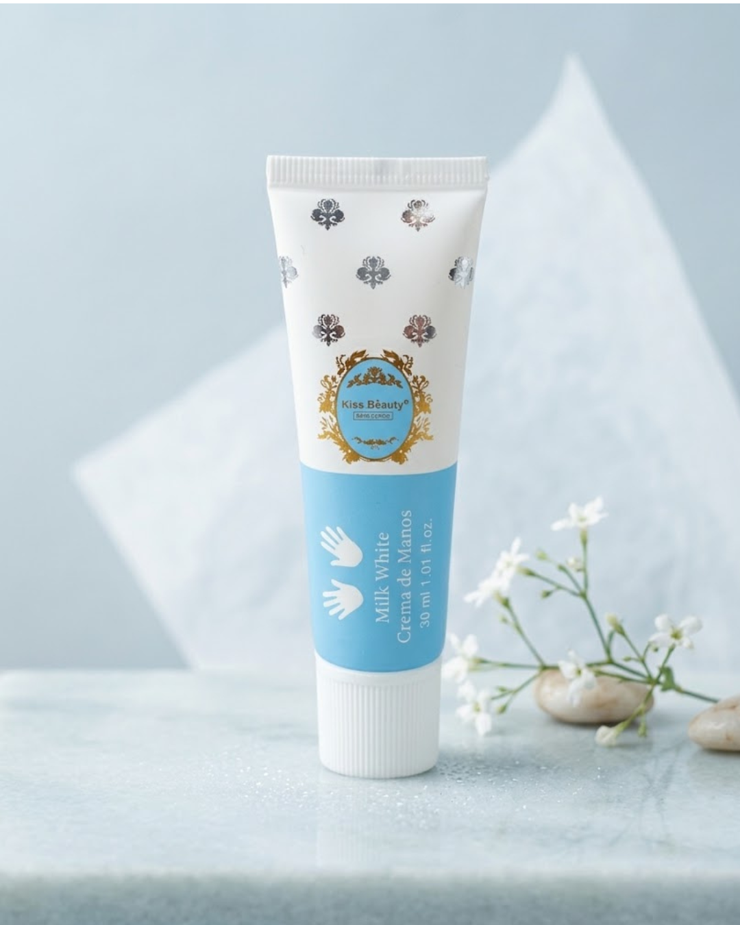 Primer plano elevado del tubo de crema de manos Kiss Beauty Milk White (Crema de Manos, 30 ml) con detalles plateados y dorados, presentado en un entorno de spa relajante con flores frescas, exfoliante y piedras de mármol.